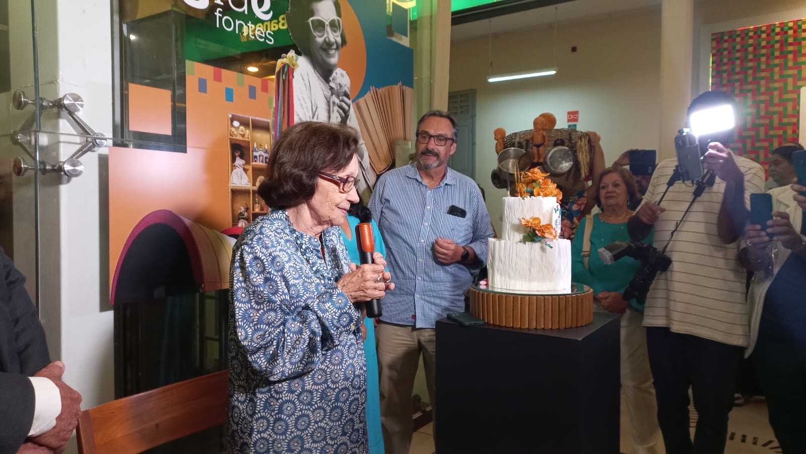 Aglaé, de vestido azul estampado e microfone na mão, diante do bolo dos seus 90 anos no Museu da Gente Sergipana